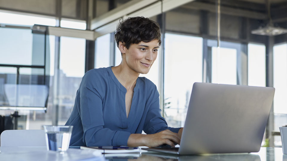 Eine Frau sitzt an einem Schreibtisch in ihrem Büro. Sie schaut während einer Onlinebesprechung auf ihren Laptop 